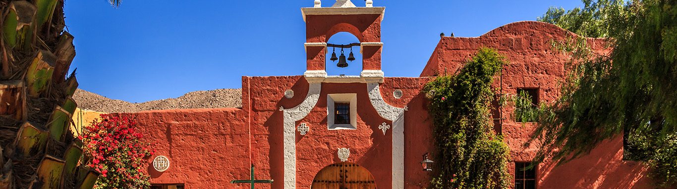 Red walls of Spanish catholic chapel with palms, trees and flowe